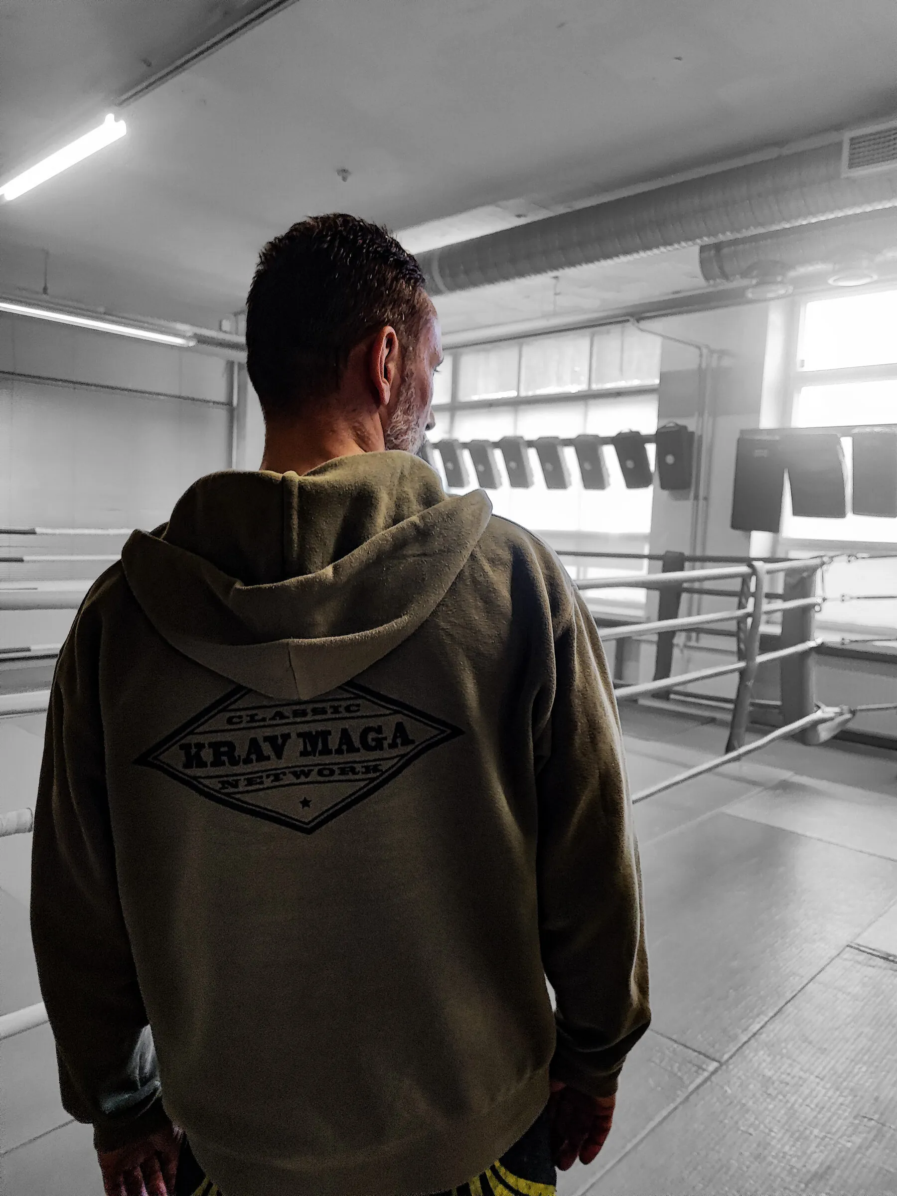 An instructor from behind in a Classic Krav Maga Network hoodie, overlooking the training hall and ring