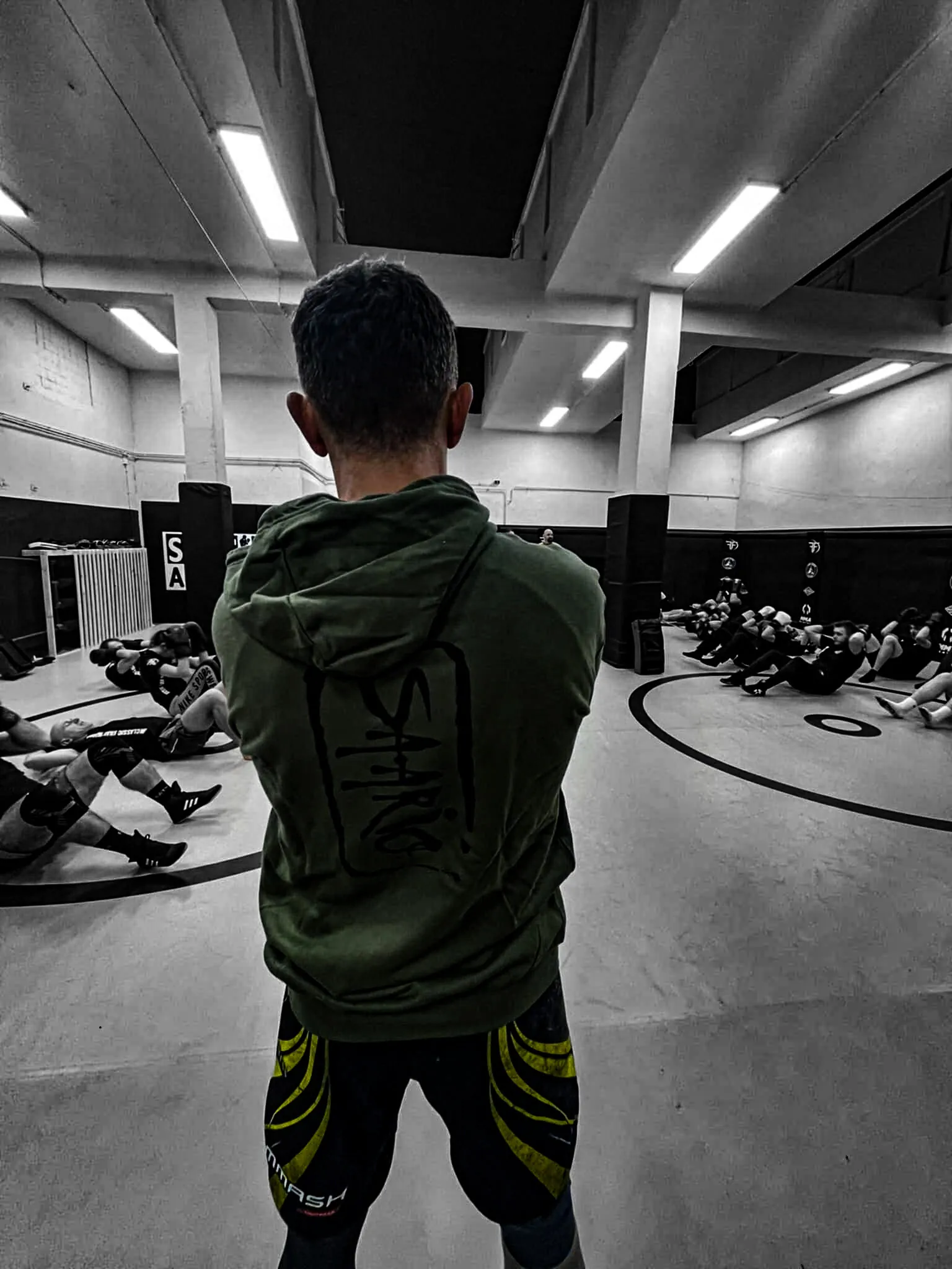 An instructor seen from behind in a Saario hoodie, quietly watching over a class working through ground drills